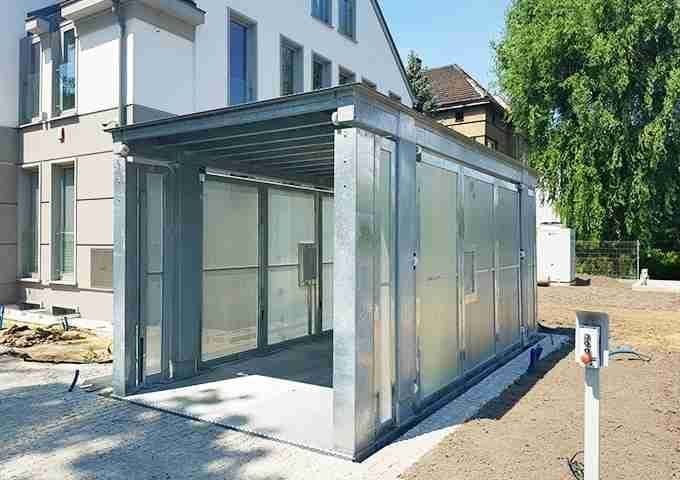 Foto reale del sistema Montauto Con Uomo A Bordo installato - struttura in vetro e metallo grigio, parcheggio esterno, edificio residenziale sullo sfondo